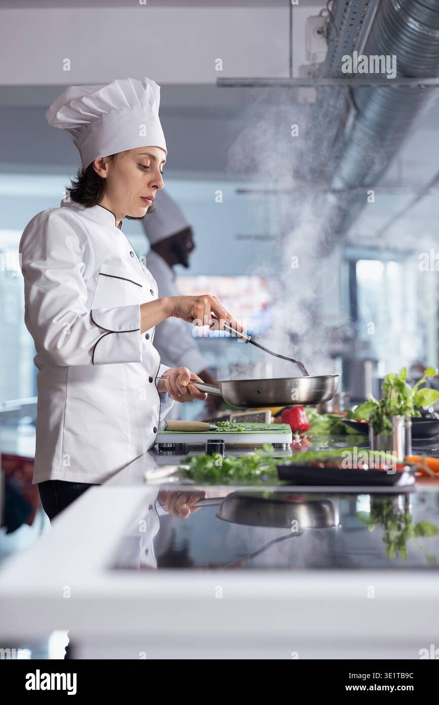 Incentrata sulla perfezione culinaria, chef donna in uniforme e cappello mescola gli ingredienti in una pentola fumante sul piano cottura, creando un piatto gourmet con precisione e passione per l'industria alimentare. Foto Stock