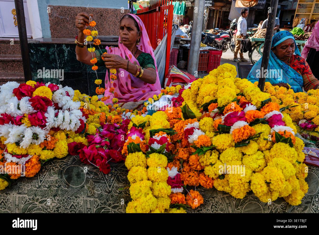 Una donna in un sari rosa assembla ghirlande di calendula in una stalla di fiori a Udaipur, con fiori colorati disposti sul tavolo. Foto Stock