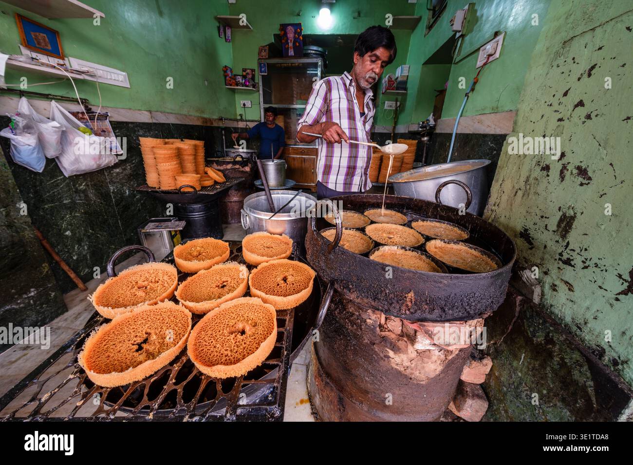 In una piccola panetteria di Udaipur, un cuoco prepara Ghevar versando l'impasto in stampi circolari su una grande stufa per creare il dolce tradizionale. Foto Stock