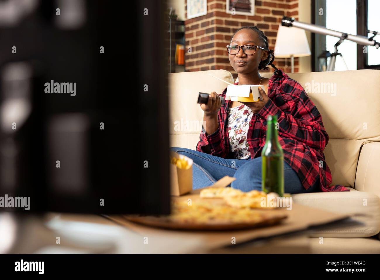 Una donna con i bicchieri si rilassa a casa, mangiando un delizioso pasto da asporto mentre sfoglia i canali TV con il telecomando. Una donna nera che si gode un pomeriggio accogliente con cibo e intrattenimento. Foto Stock