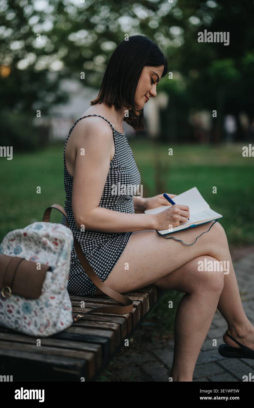 Donna che scrive su un notebook sulla panca del parco indossando un abito estivo a scacchi e uno zaino Foto Stock