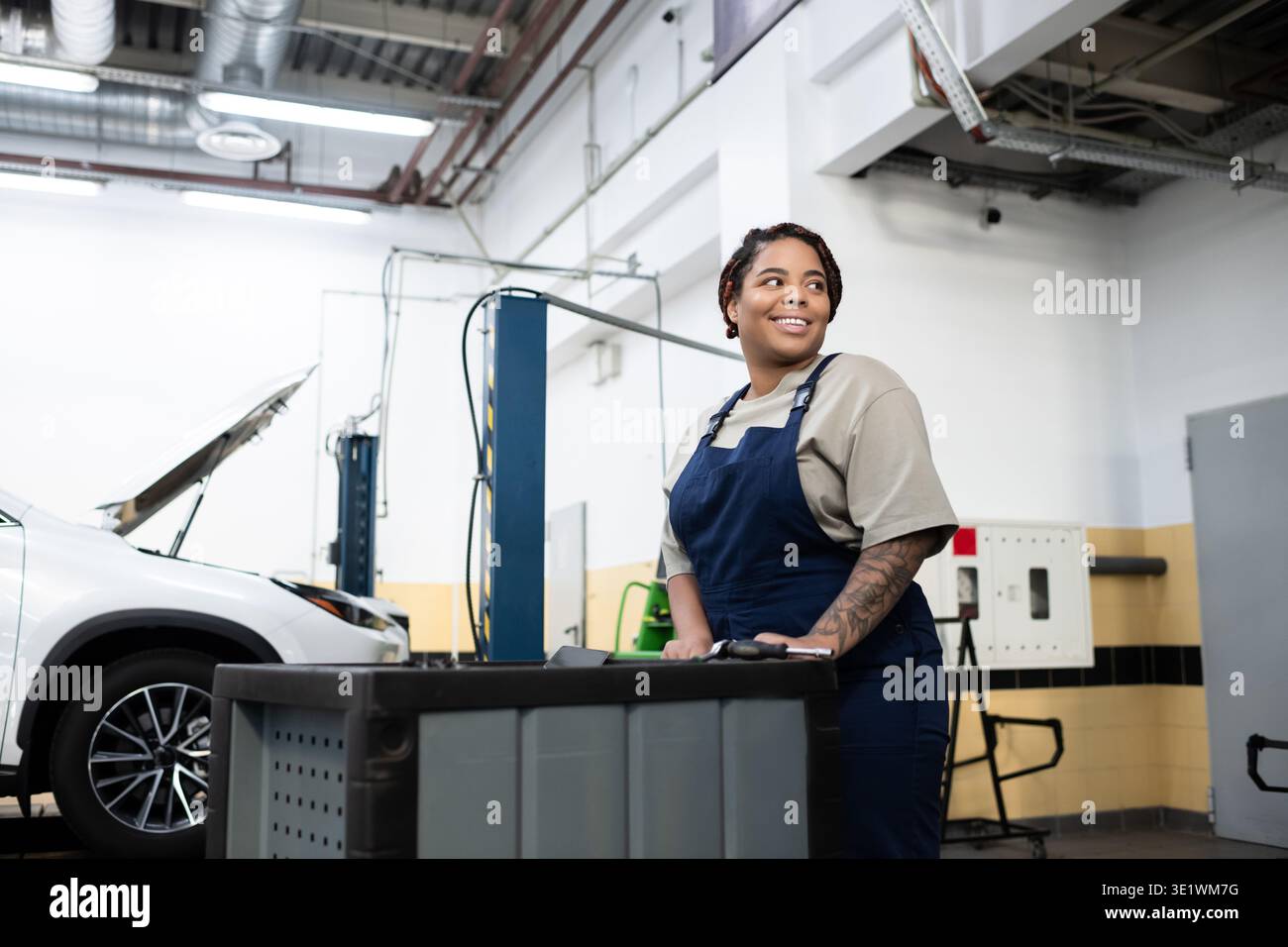 Una giovane donna esperta in tuta sorride mentre maneggia gli attrezzi in un'officina di riparazione auto molto affollata. Foto Stock