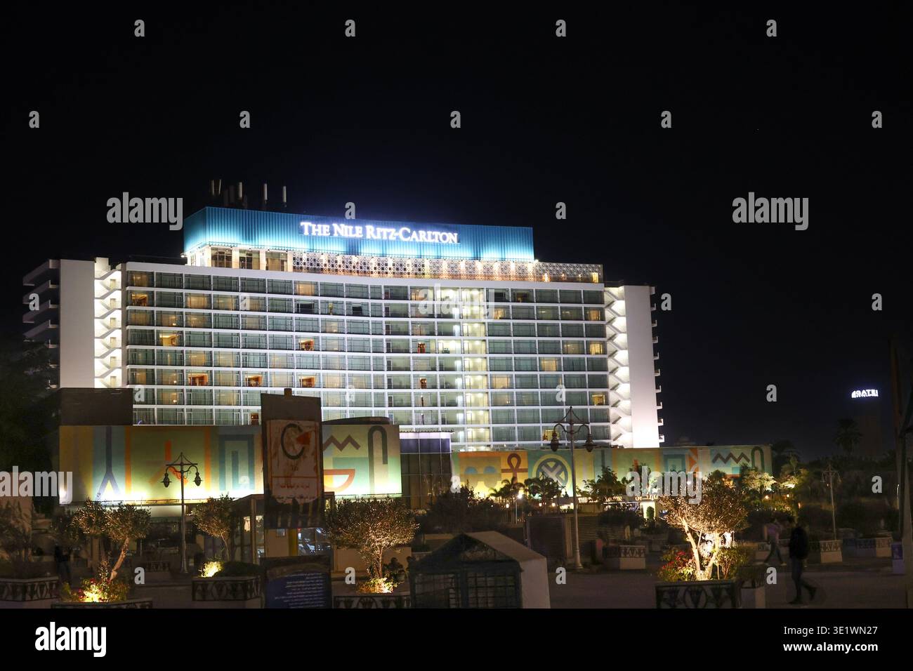 Vista notturna dell'hotel Nile Ritz-Carlton nel centro del Cairo, in Egitto, con la sua facciata illuminata a più piani, le palme in primo piano e un vib Foto Stock