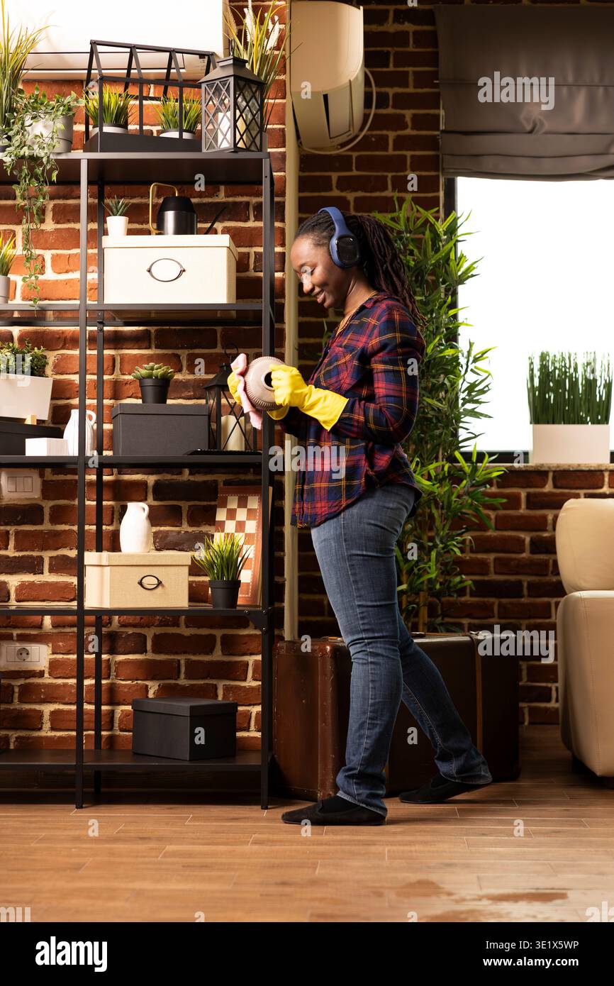 La casalinga afroamericana sorridente si trova vicino agli scaffali, pulisce il vaso con un panno mentre ascolta la musica. Donna nera che indossa cuffie e guanti, ama svolgere le faccende domestiche in casa. Foto Stock