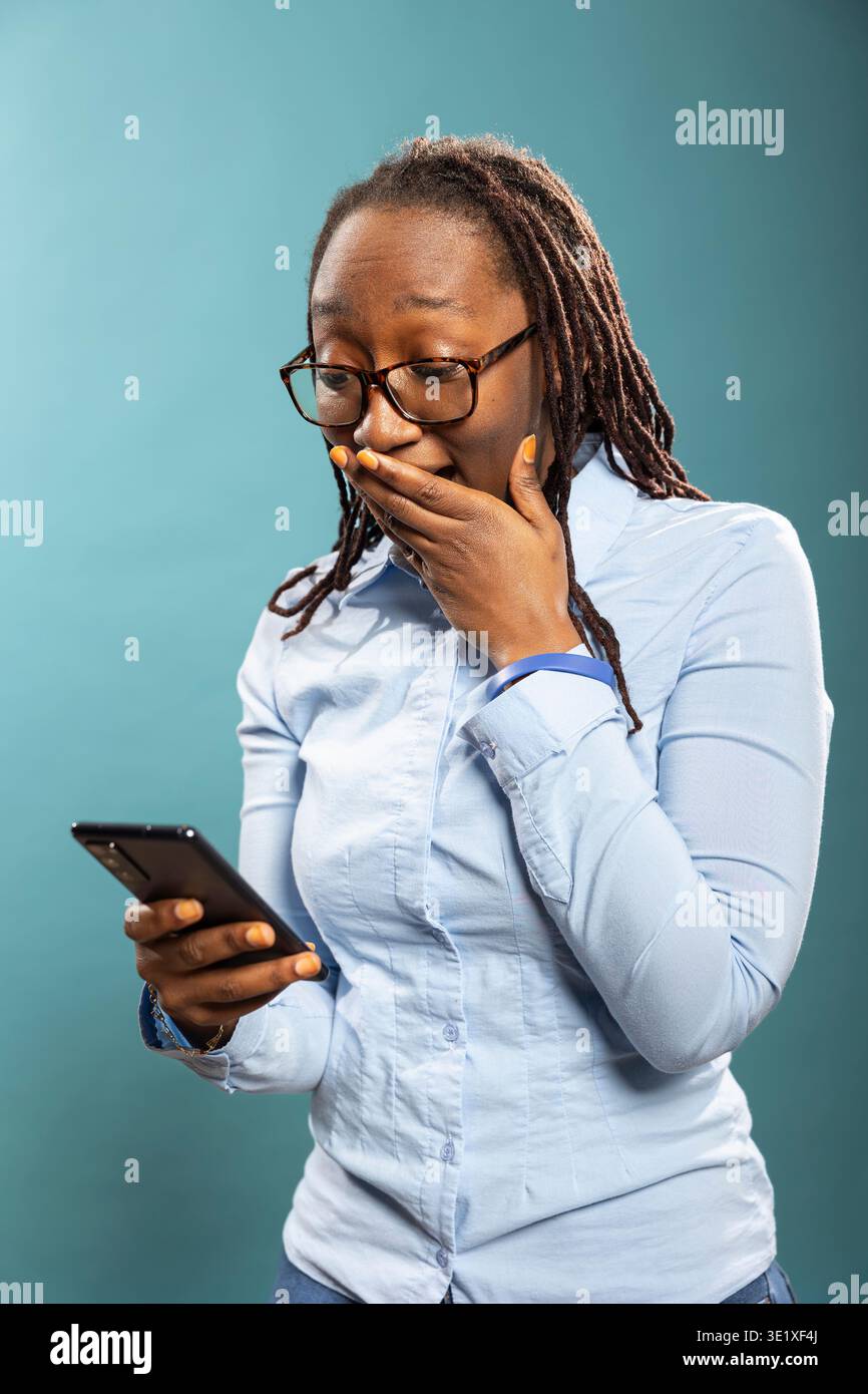 La giovane donna sbalordita fissa il suo smartphone, coprendo la bocca con stupore dopo aver ricevuto notizie meravigliose. Sorpresa afroamericana che controlla il suo dispositivo mobile sotto shock. Foto Stock