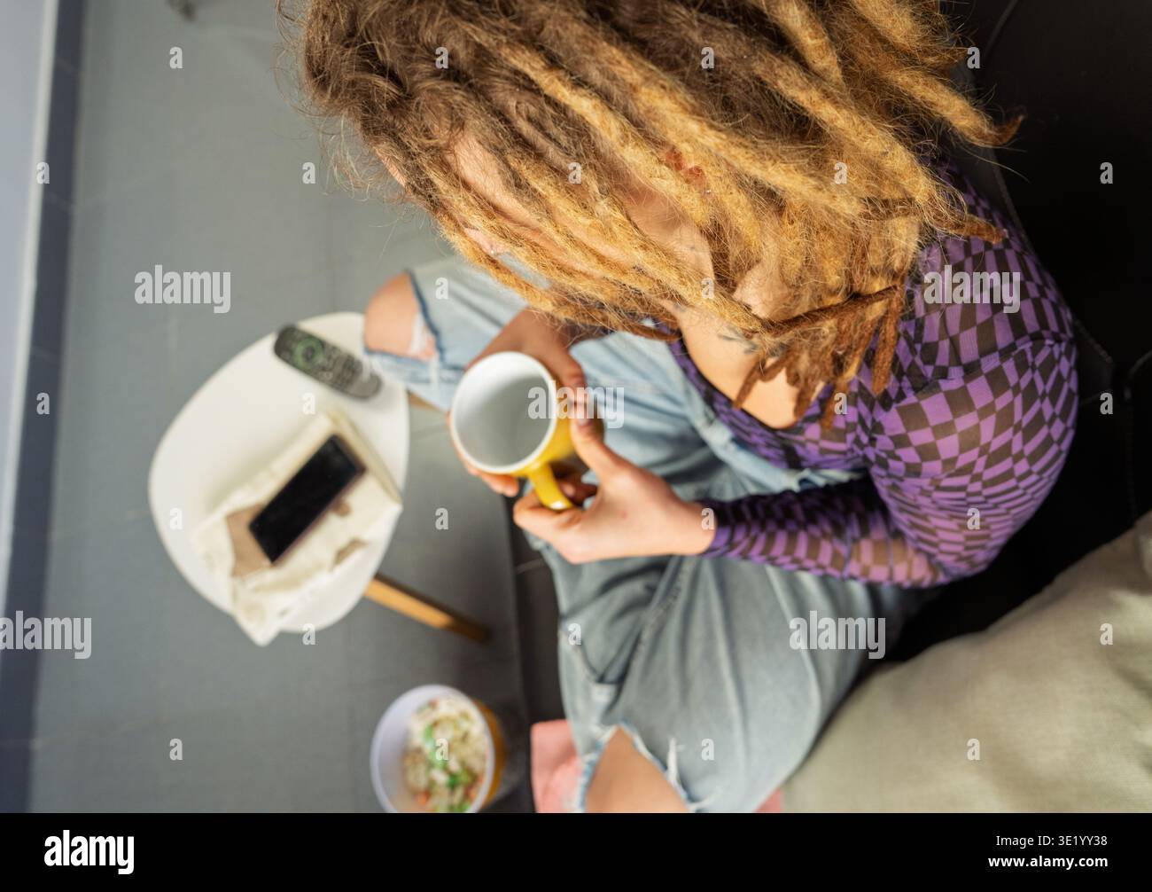Persona con dreadlock che tiene in mano una tazza gialla, rilassandosi in un ambiente informale a casa Foto Stock