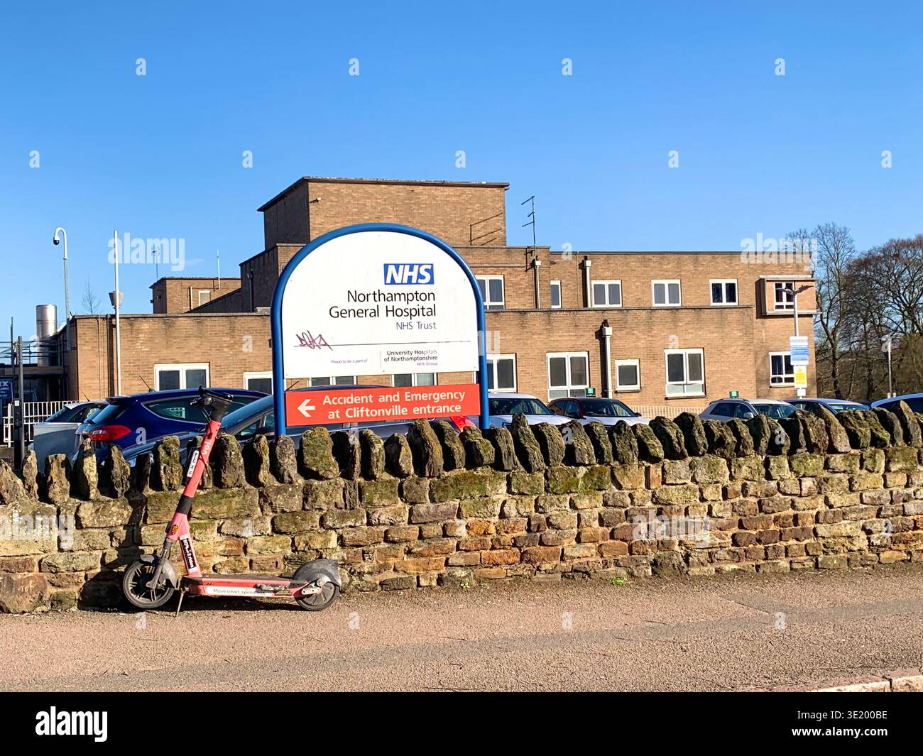 Ingresso principale del Northampton General Hospital in una giornata di sole con veicoli parcheggiati e uno scooter elettrico fuori dal muro di pietra di confine un cartello dirige - Immagine stock catturata con smartphone