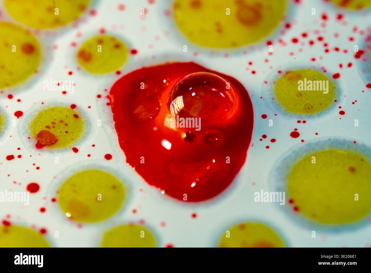 Gocce colorate di liquidi gialli e rossi si trovano su una superficie bianca liscia. La goccia rossa è più grande e circondata da piccoli puntini gialli in un setti luminoso Foto Stock