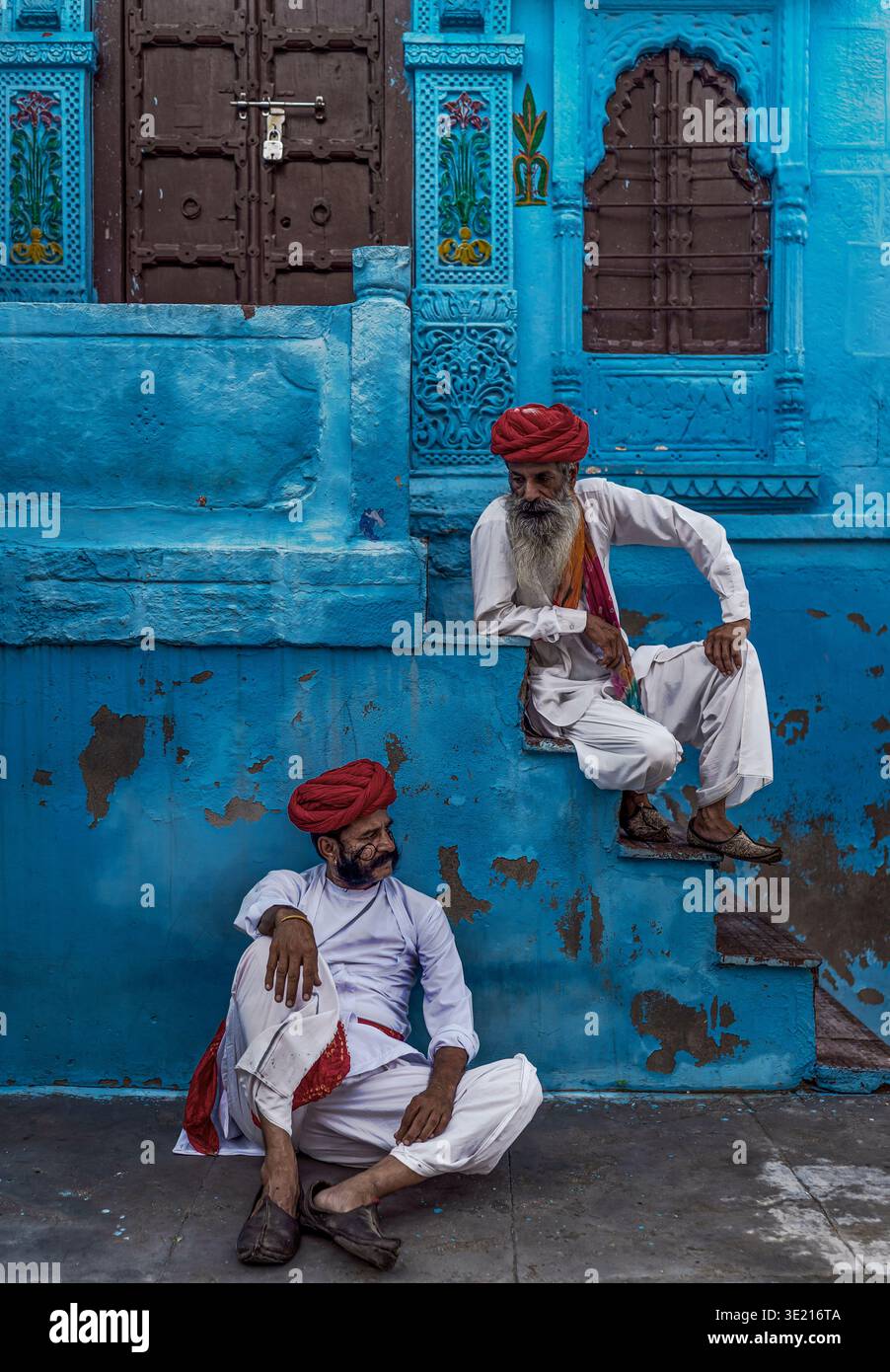 Due uomini in abbigliamento tradizionale seduti contro il muro blu nel Rajasthan, in India Foto Stock