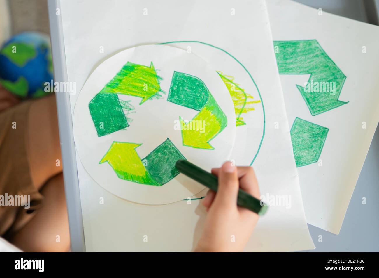 Vista ad angolo alto della mano del bambino con simbolo di riciclaggio colorato e pastello verde a casa. Riciclo e un concetto di vita ecocompatibile. Foto Stock