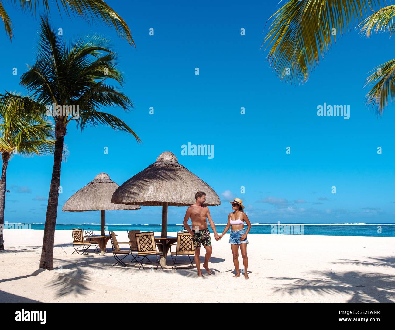 Su una splendida spiaggia di Mauritius, una coppia cammina mano nella mano, circondato da palme ondeggianti e ombrelloni di paglia. Foto Stock