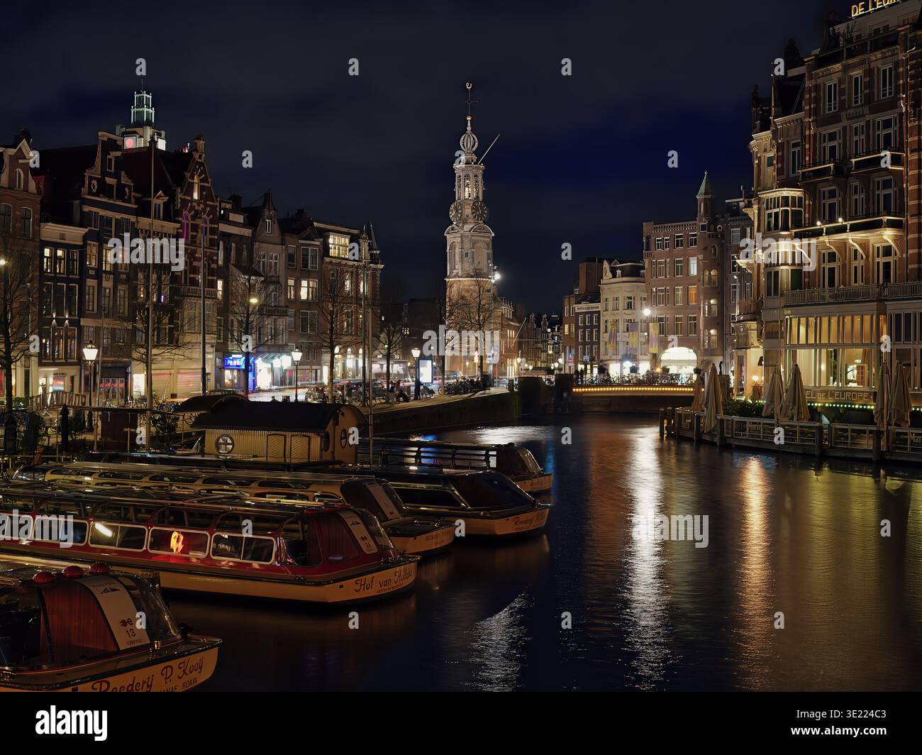 La torre dell'orologio Muntorren, alta contro il cielo notturno, con gli edifici sul canale illuminati di notte, dove si incontrano i canali Amstel e Singel. Foto Stock