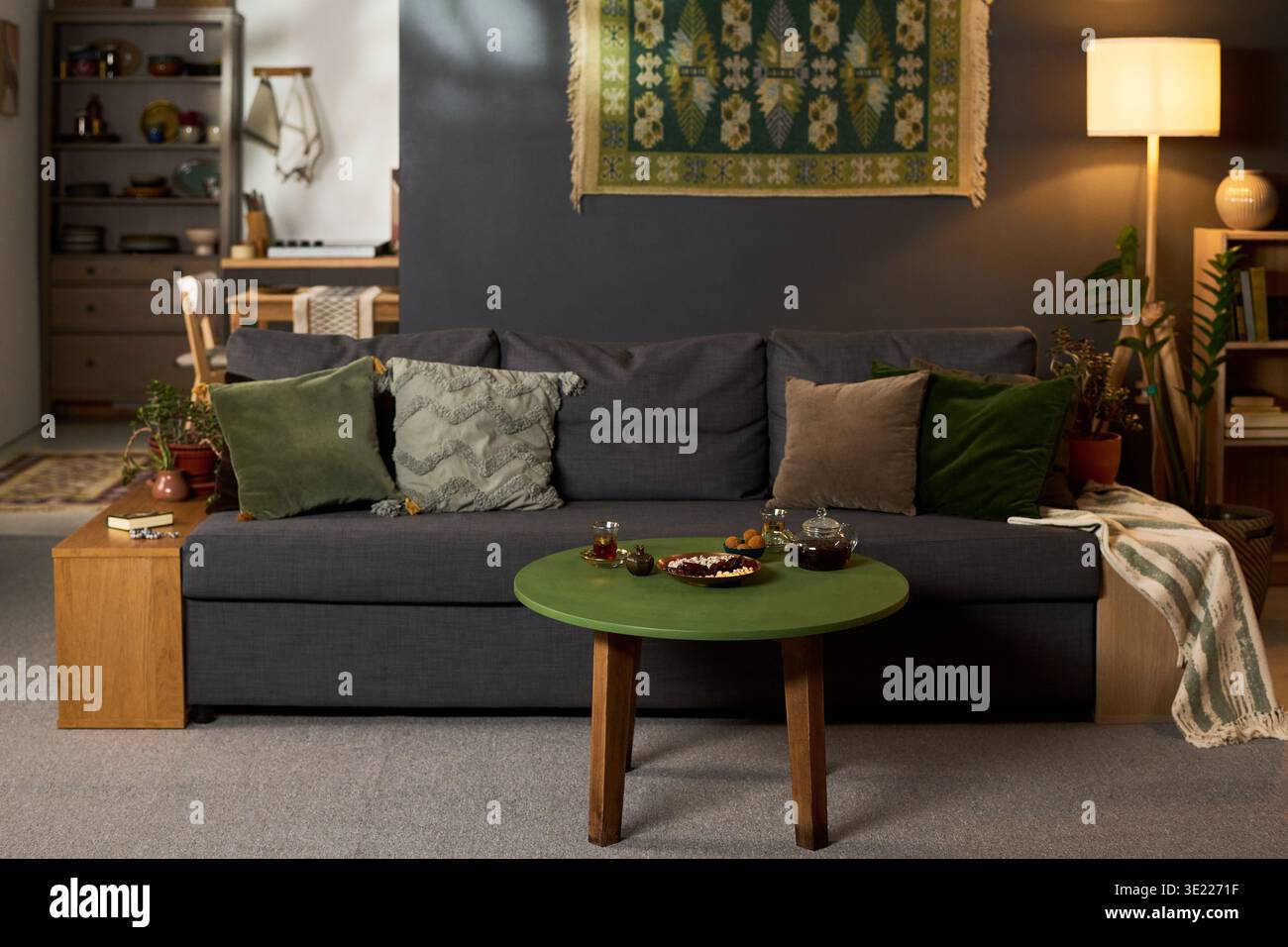 Salotto con tavolino rotondo verde con set da tè e spuntini davanti al divano grigio con cuscini multipli, sfondo con libreria, lampada e pensile decorativo Foto Stock