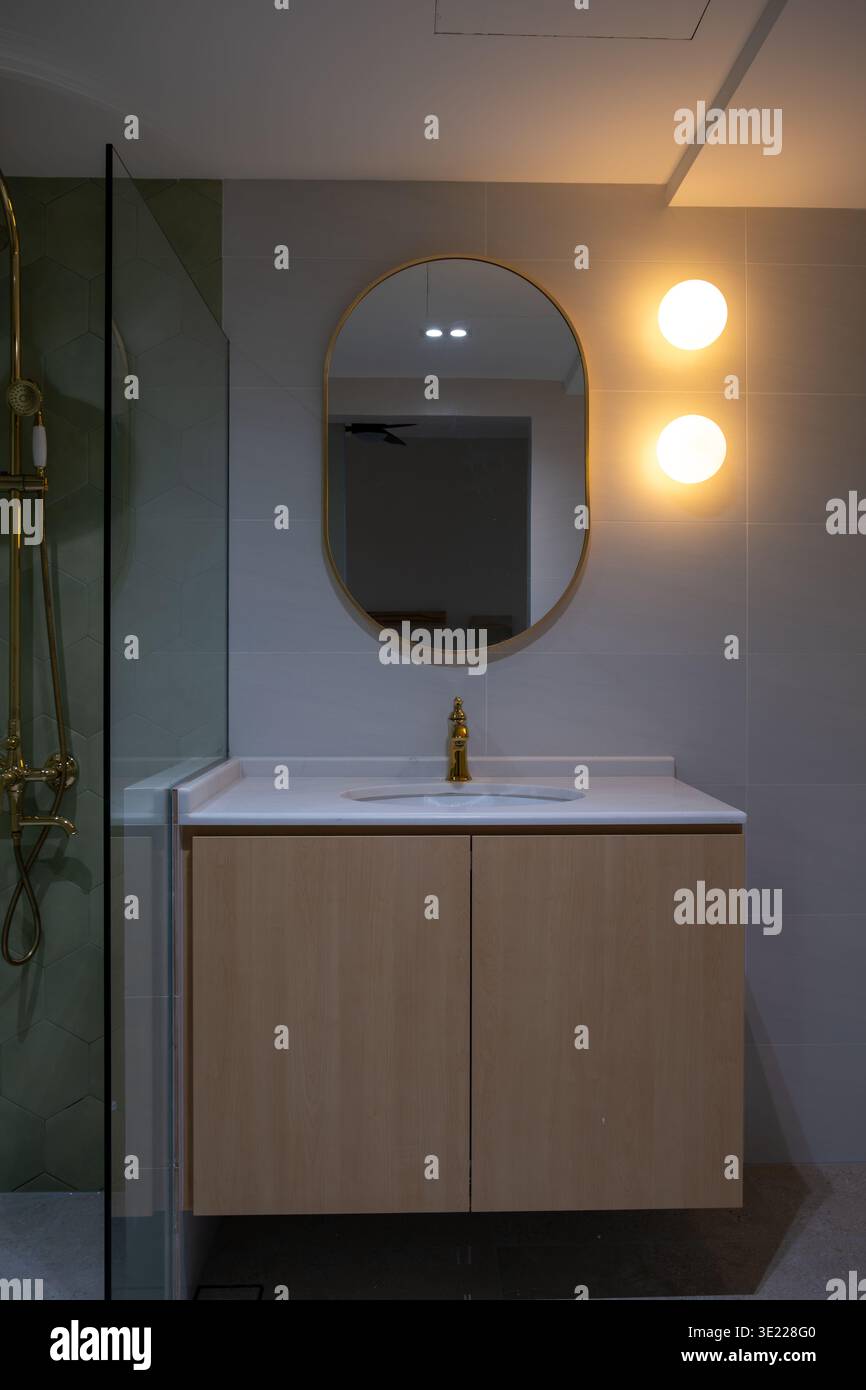 Bagno moderno con specchio rotondo e luce calda. Il lavandino in legno e il rubinetto dorato aggiungono eleganza. Il box doccia in vetro riflette il bagliore dell'ambiente. Pulizia, Foto Stock