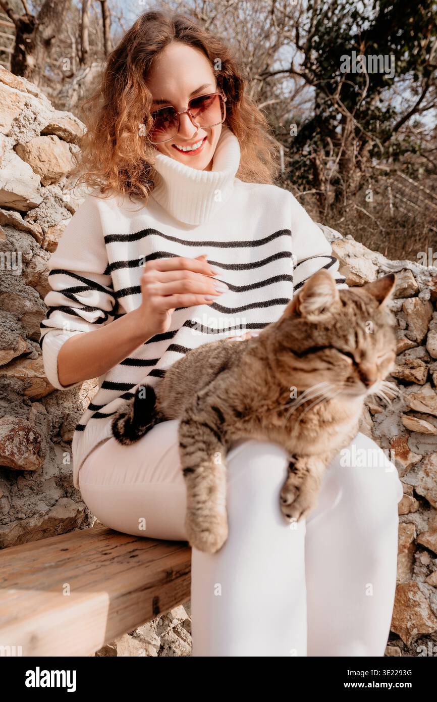 Gatto donna che accarezza la ragazza sorridente con un maglione a righe si rilassa all'aperto mentre si siede su una panchina con il suo animale domestico Foto Stock