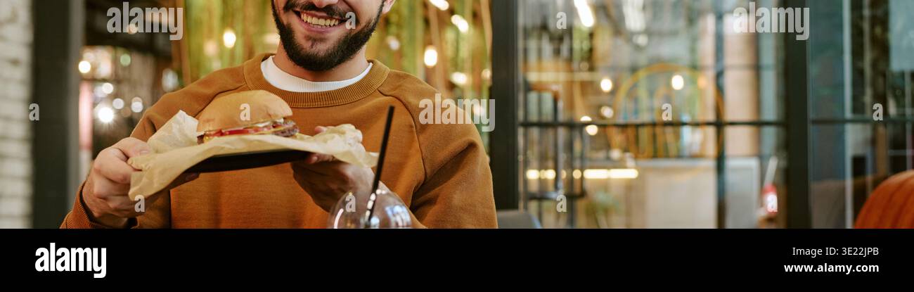 Giovane adulto che sorride mentre tiene in mano un hamburger nel ristorante moderno Foto Stock