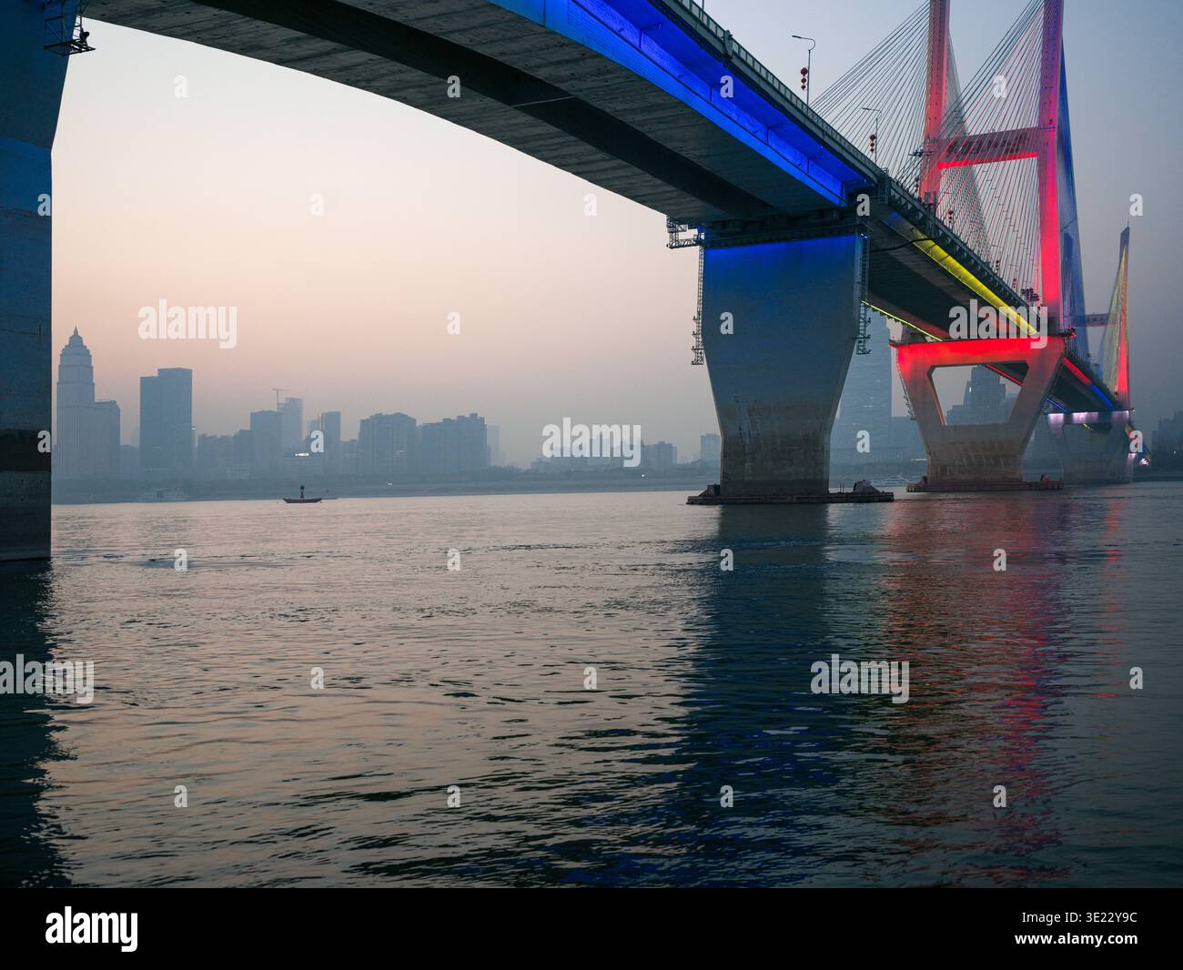 Ampia vista di un grande ponte sospeso che attraversa un ampio fiume, splendidamente illuminato con luci LED rosse e blu contro un cielo crepuscolo. T Foto Stock