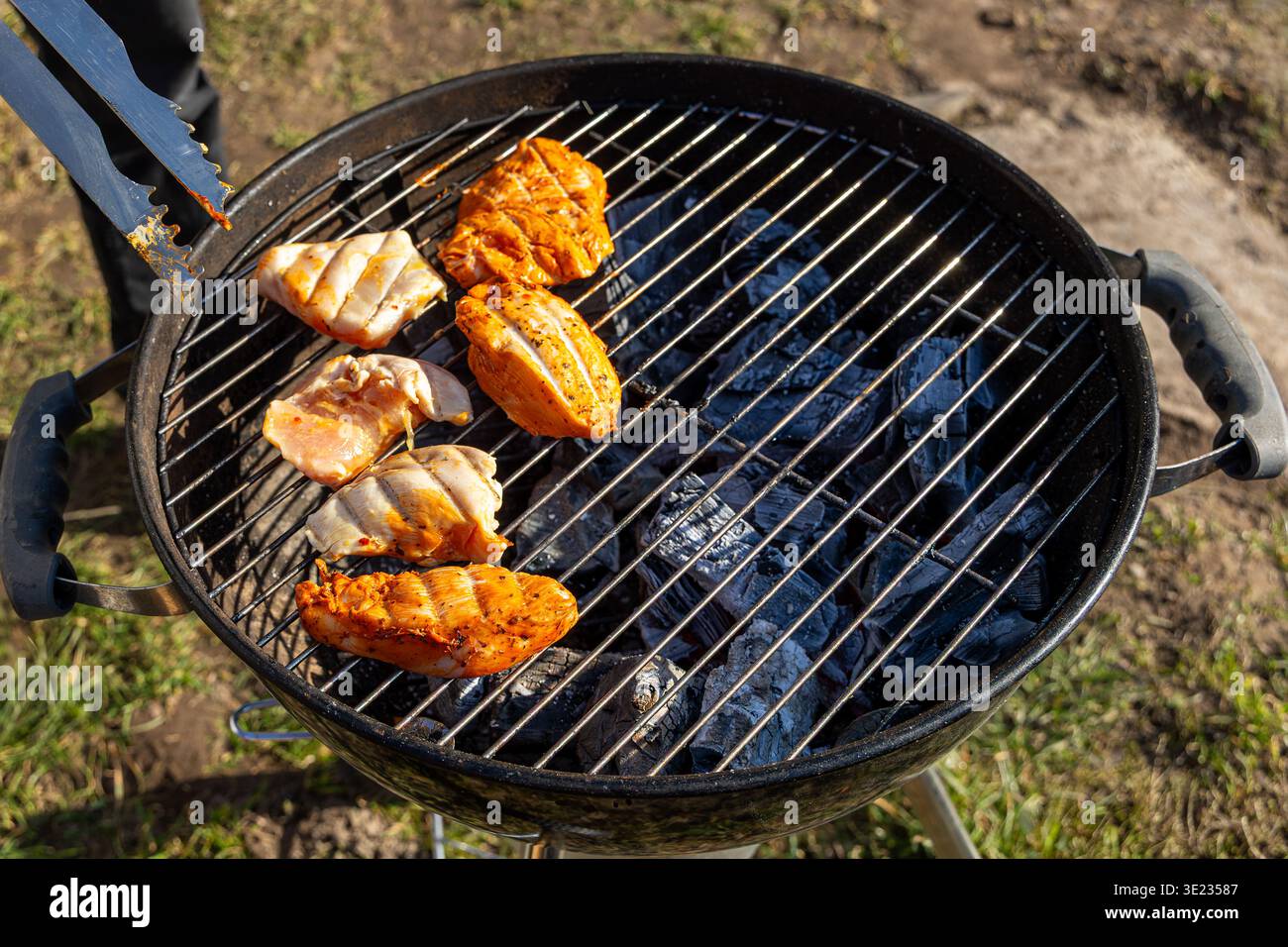 Filetti di pollo grigliati su barbecue a carbone. Primo piano di filetti di pollo marinati in cottura su una griglia a carbone all'aperto, con pinze metalliche e carboni caldi Foto Stock