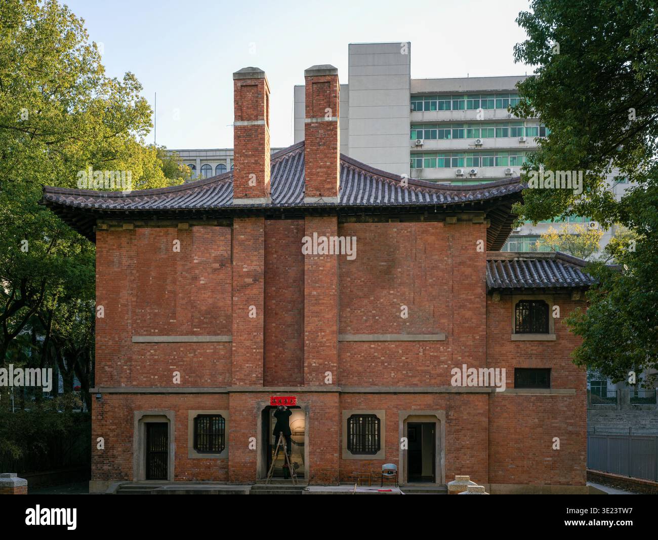 Un edificio storico simmetrico in mattoni rossi caratterizzato da due alti camini centrali e da un tetto tradizionale, situato tra alberi verdi con edifici moderni Foto Stock