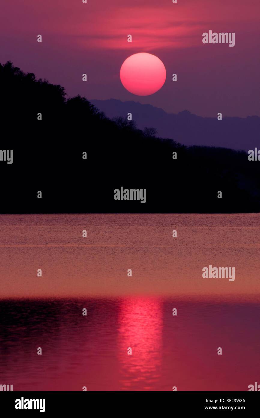 Un drammatico tramonto cremisi su un lago calmo con un sole rosso brillante che si riflette sull'acqua, incorniciato da sagome di montagna scure. La scena crea un po Foto Stock