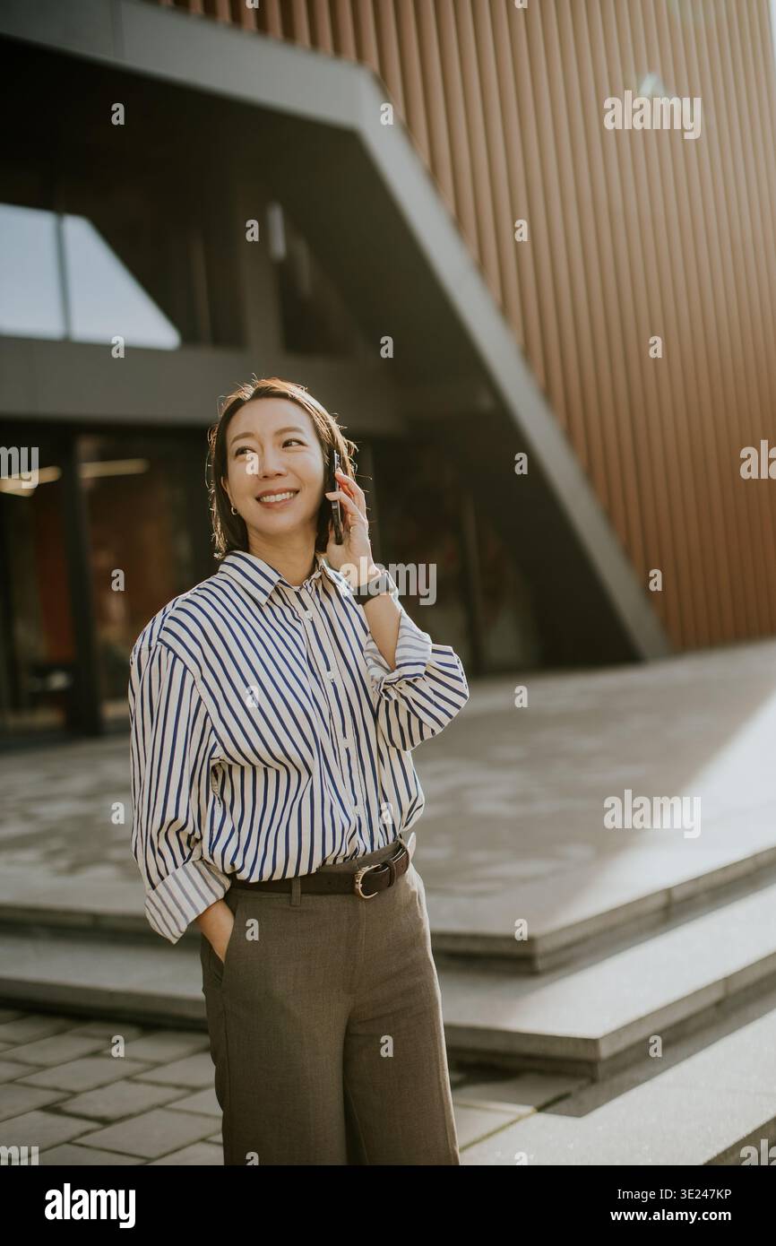 Una donna coreana si trova fuori da un edificio moderno. Sta sorridendo e parlando al telefono. Il sole splende su di lei mentre le piace la conversazione. Foto Stock