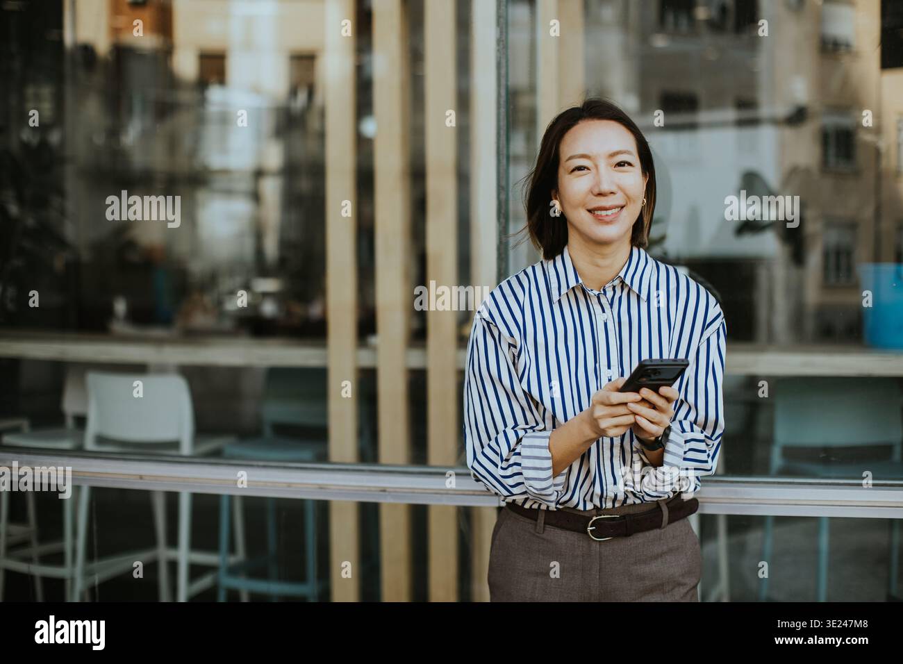 Una donna coreana sorride mentre tiene il suo smartphone. Si appoggia contro una ringhiera all'esterno di un edificio moderno. La scena la mostra in un abbigliamento casual Foto Stock