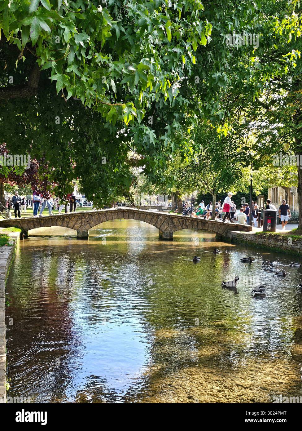 Una vista ravvicinata di un ponte di pietra sul fiume Windrush a Bourton-on-the-Water mostra folle che si godono una giornata intensa in tarda estate in questo villaggio delle Cotswolds. - Immagine stock catturata con smartphone