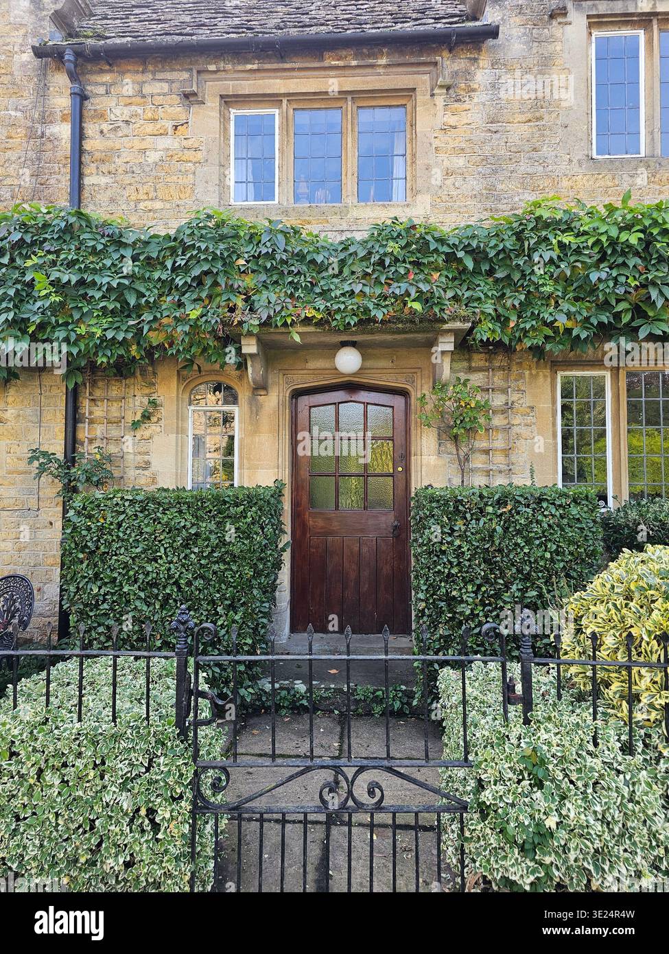 Affascinante cottage Bourton-on-the-Water con cancello di ferro nero, topiario pulito, pareti coperte di fogliame e sentiero che conduce ad una porta d'ingresso marrone - Immagine stock catturata con smartphone
