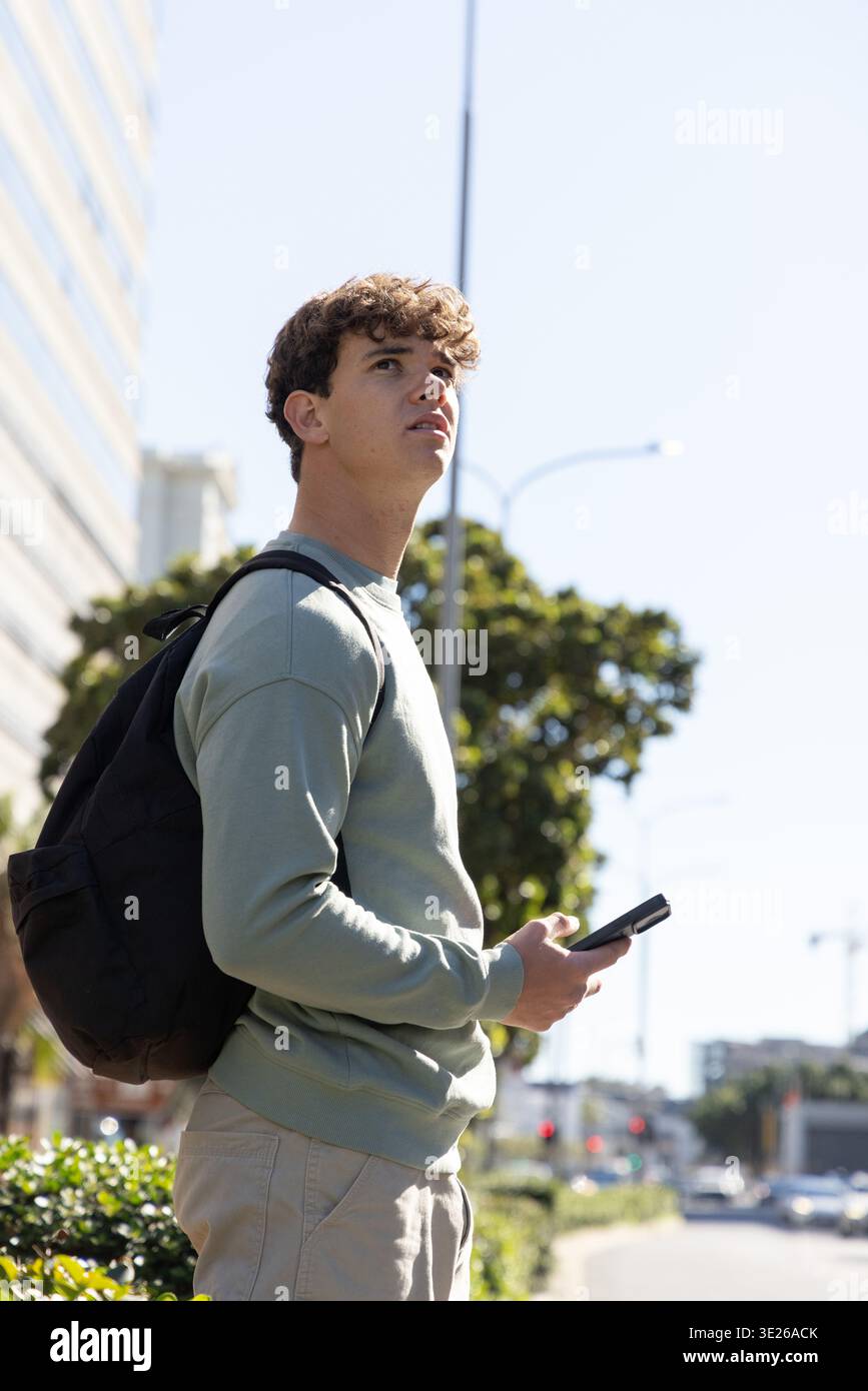 Adolescente in piedi sul marciapiede della città tenendo il telefono, indossando una felpa verde chiaro e uno zaino Foto Stock