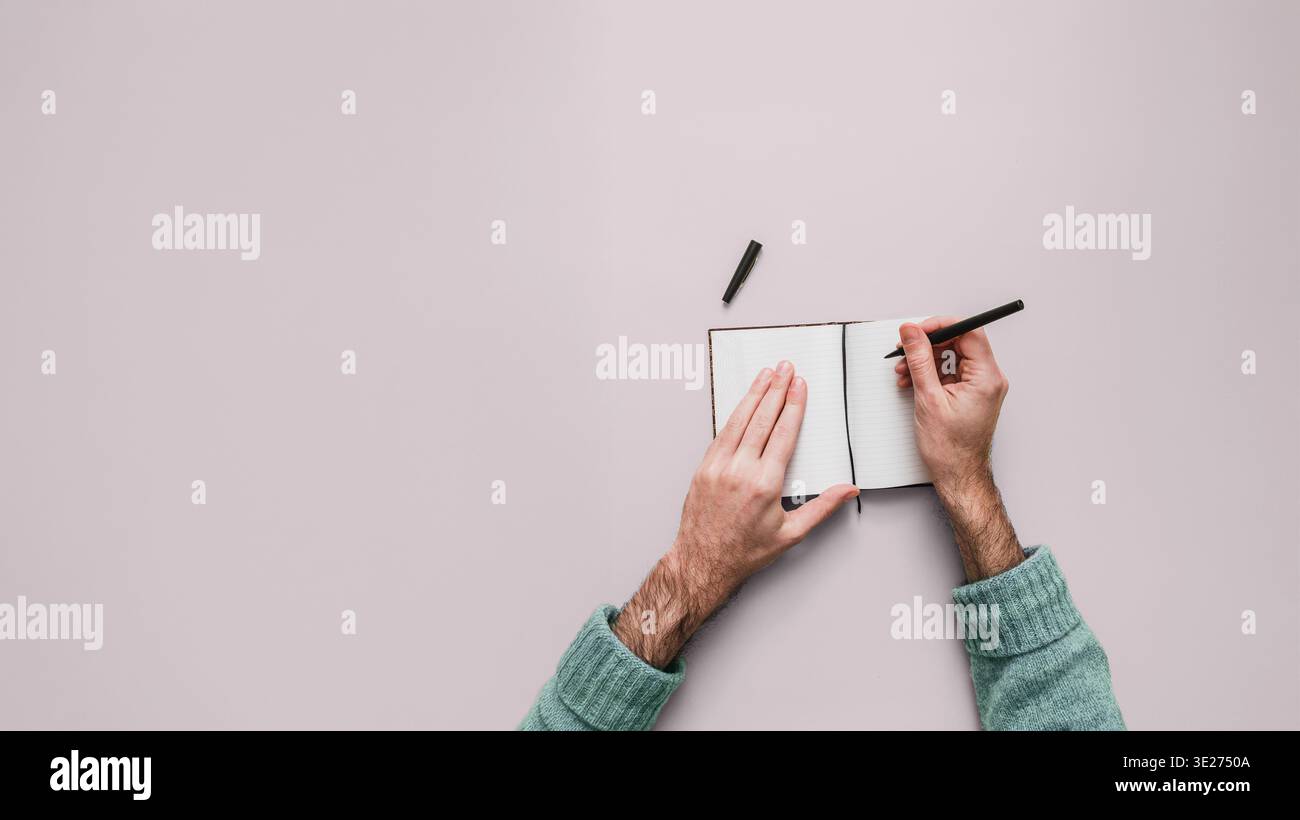 Mani maschili che scrivono in un piccolo quaderno aperto con penna nera. Sfondo lilla uniforme con ampio spazio di copia a sinistra. Seconda penna visibile. Sovratesta V Foto Stock