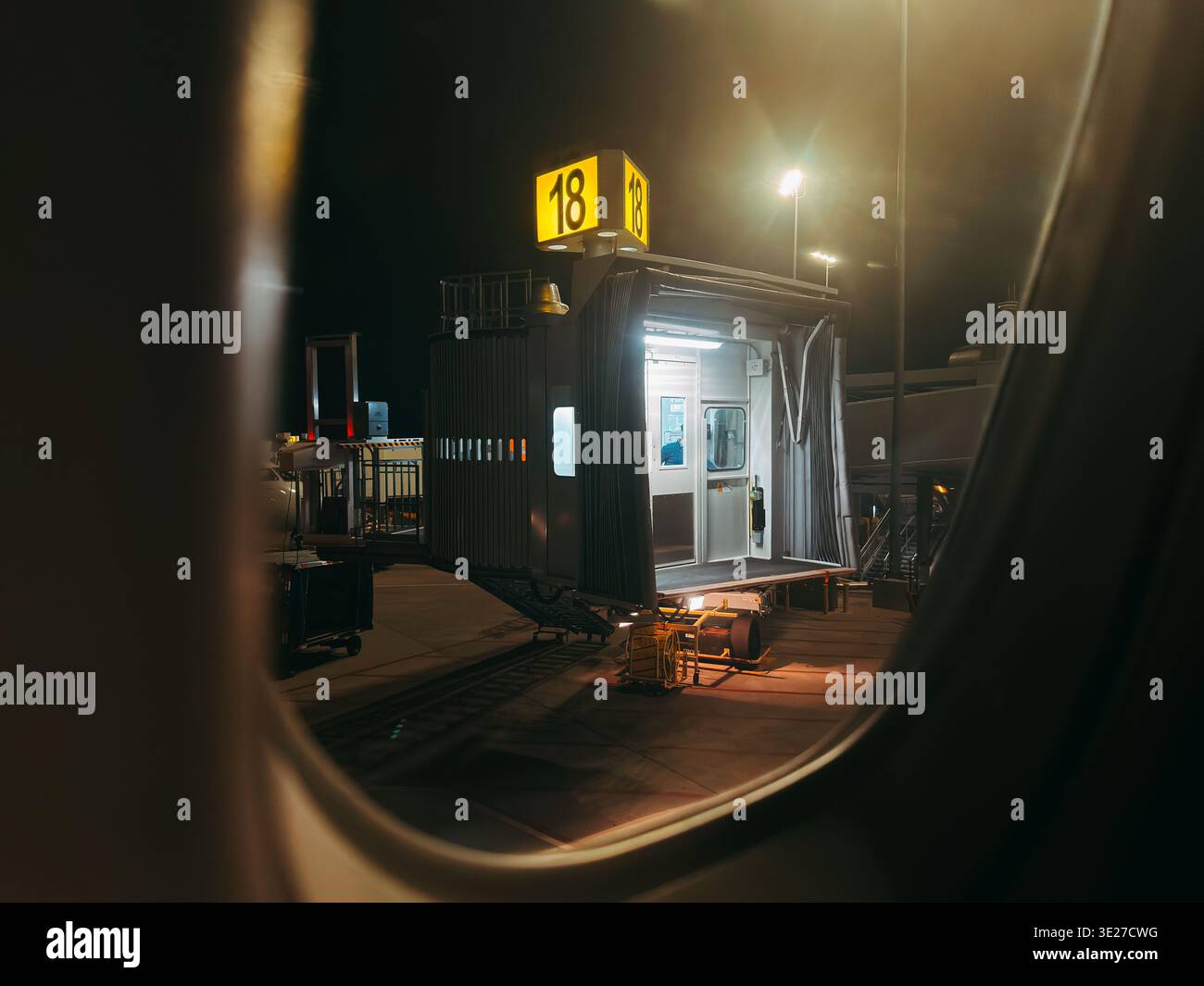 Vista notturna dal finestrino dell'aereo del cancello 18 illuminato del ponte jet, con attrezzature per terminal e rampa Foto Stock