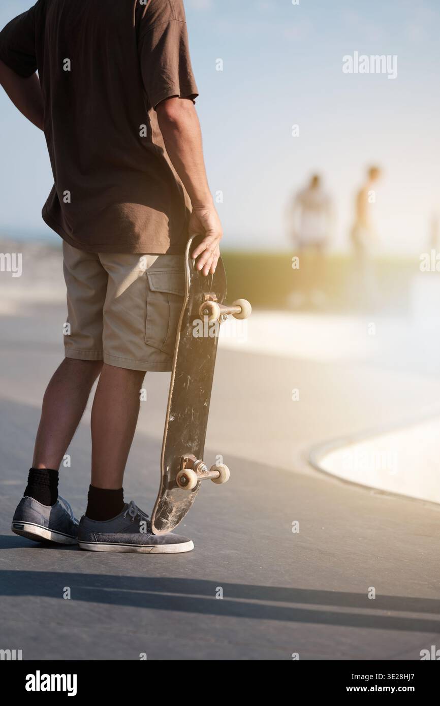 Giovane skateboarder che porta una tavola in uno skatepark di cemento durante l'ora d'oro Foto Stock