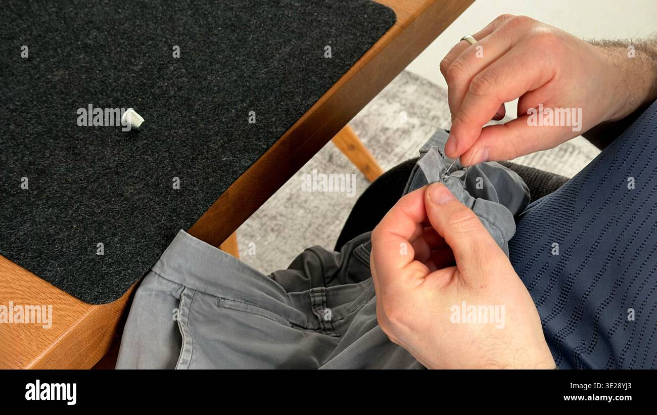 Uomo che cucisce bottone su tessuto grigio della camicia mentre è seduto a un tavolo di legno con superficie scura in feltro Foto Stock
