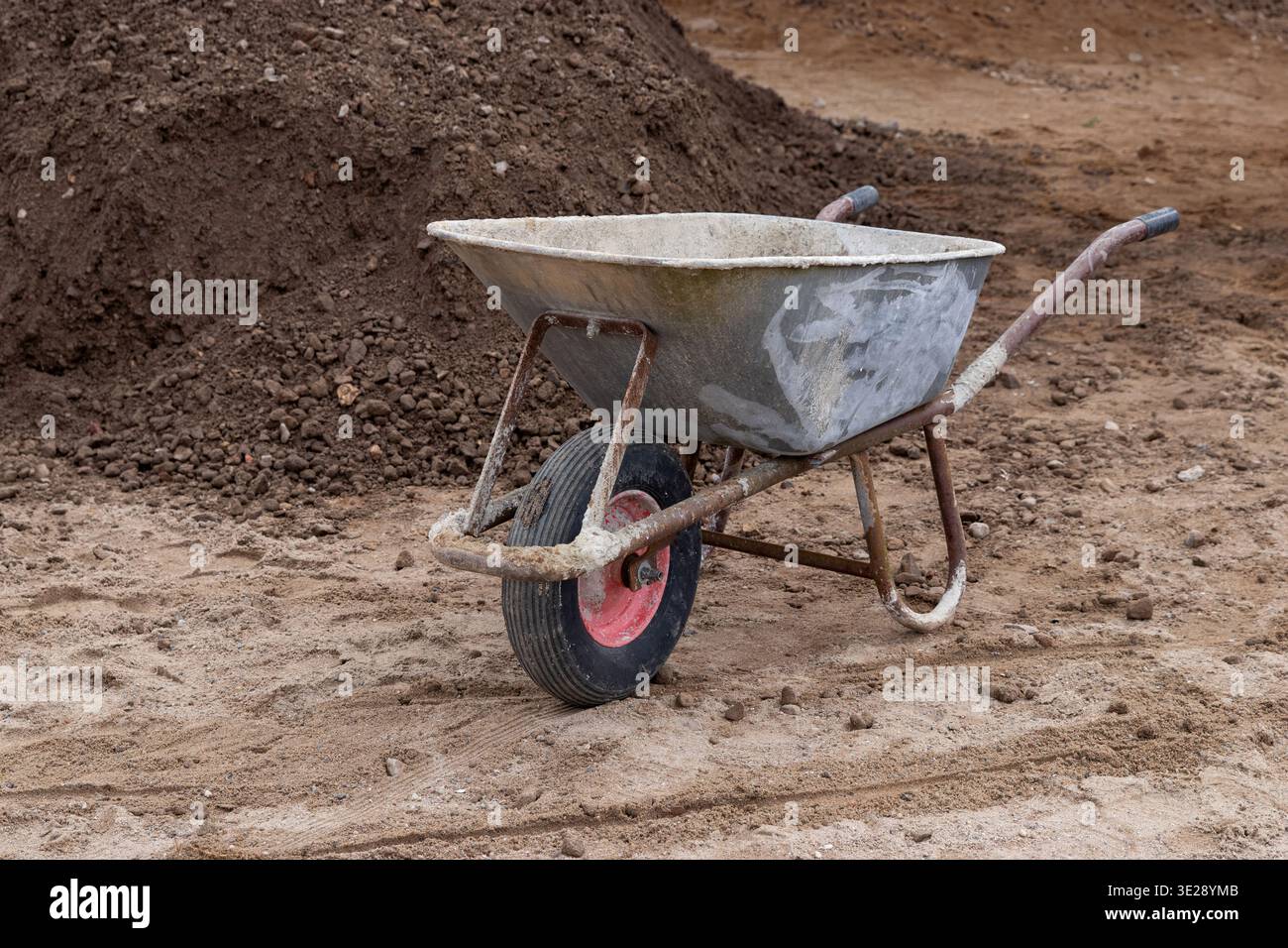 Carriola in metallo con una ruota singola posizionata su un terreno sabbioso vicino a un grande mucchio di sporcizia in un cantiere edile Foto Stock