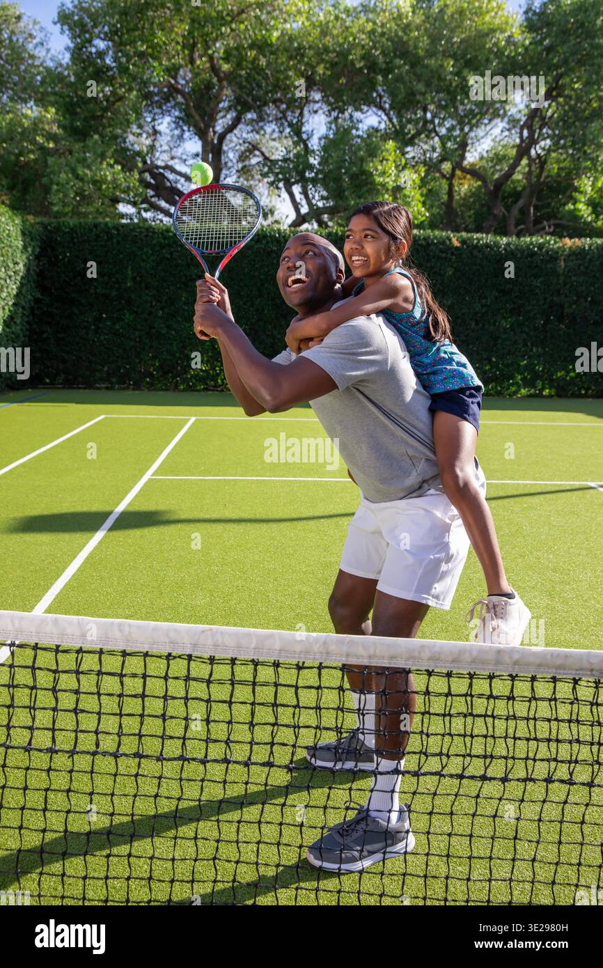 Papà afroamericano che porta la figlia sulla schiena e pratica il tennis con la racchetta sul campo. Sport, famiglia, outdoor, tempo libero, dinamica, attivo, connessione || modello rilasciato Foto Stock