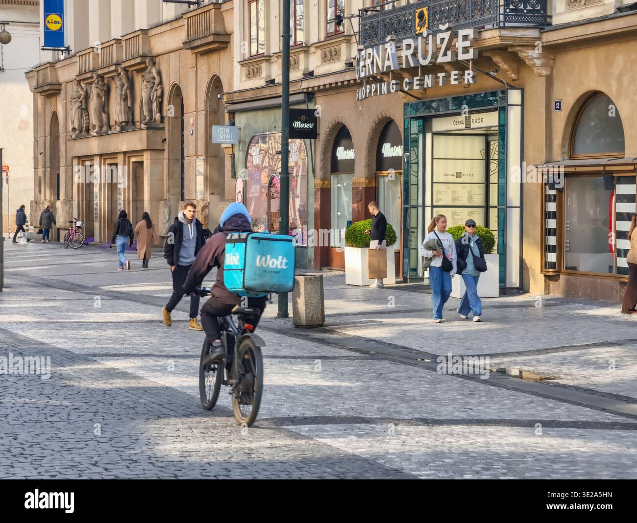 Praga, Repubblica Ceca - 19 marzo 2026: Corriere a domicilio in bicicletta nel centro storico di Praga. Corriere che attraversa la strada pedonale acciottolata Foto Stock