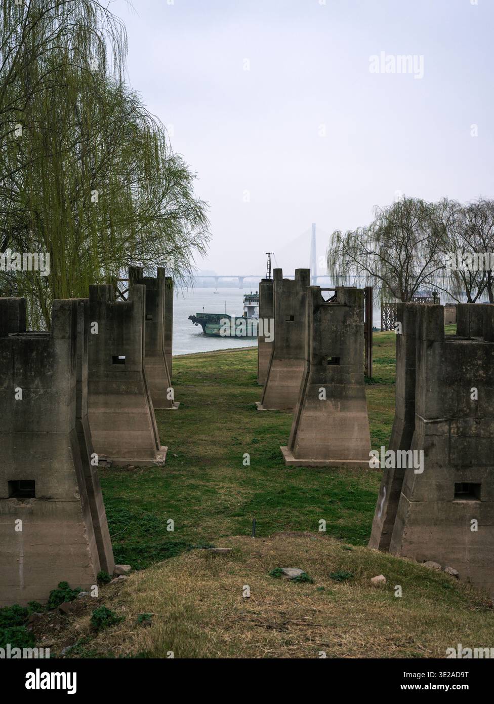I pilastri di cemento intempestivo di una struttura industriale abbandonata si trovano su una sponda erbosa del fiume, con una nave da carico verde che passa sull'acqua dietro. Ill Foto Stock