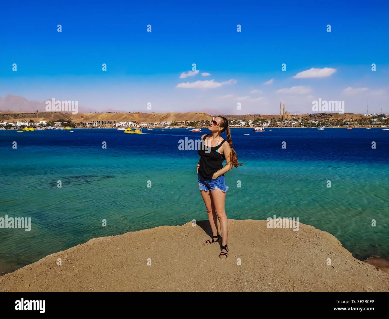 Una giovane donna adulta con i capelli intrecciati si trova sul bordo di una scogliera sul Mar Rosso a Sharm El Sheikh, Egitto. Una bella e snella posa di un turista europeo Foto Stock