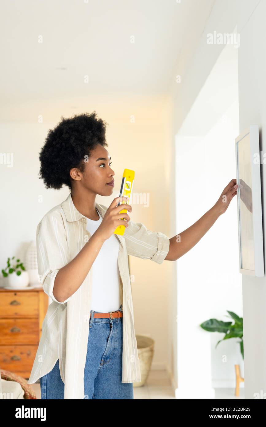 Con una livella a bolla gialla, donna che regola le opere d'arte incorniciate su una parete bianca a casa, indossa jeans. Interni, minimalisti, luminosi, arredi, piante, legno, Foto Stock