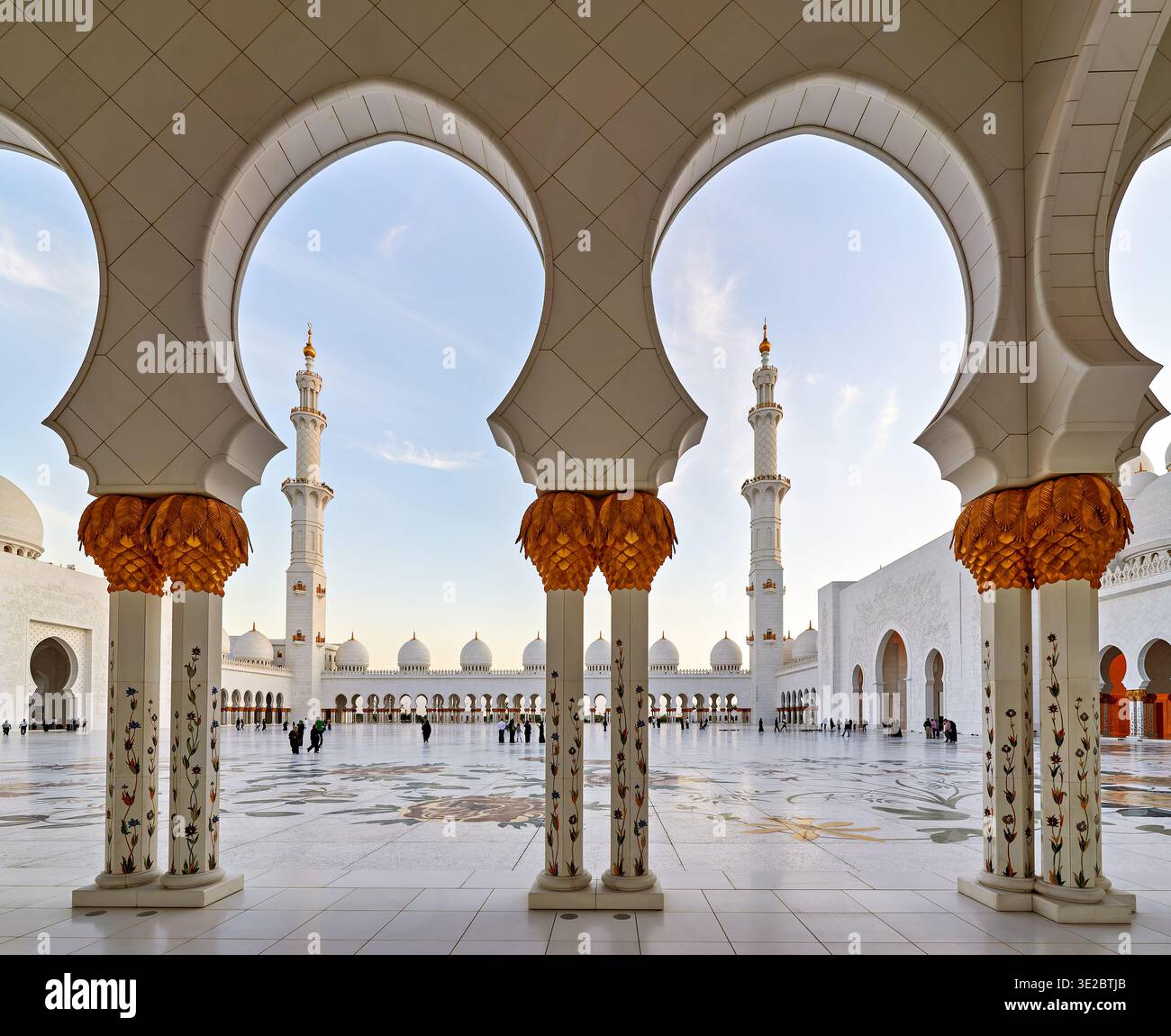 La gente cammina attraverso il cortile della grande Moschea dello Sceicco Zayed ad Abu Dhabi durante il tramonto. L'architettura unica si erge intorno a loro. Foto Stock