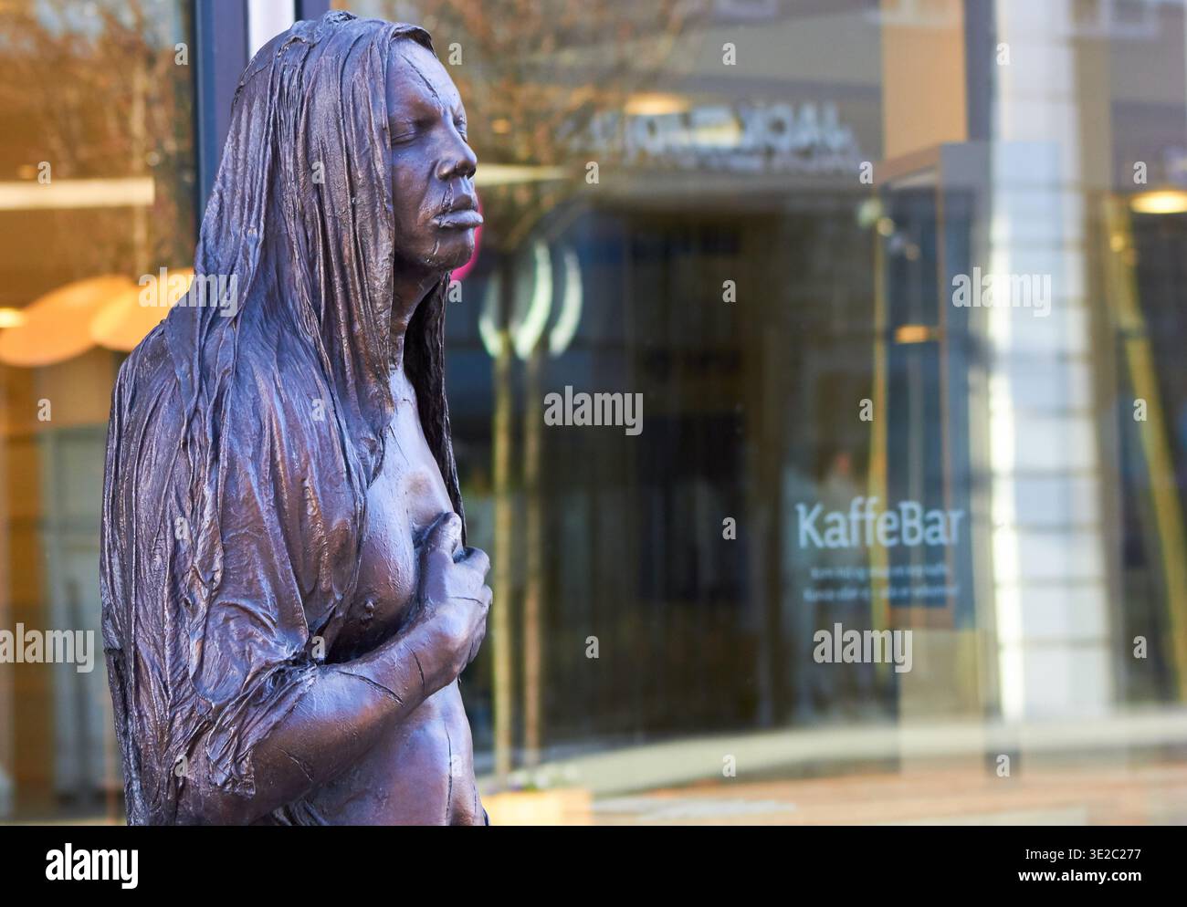 Una dettagliata scultura in bronzo di una donna con lunghi capelli parzialmente coperti dal viso si trova in un contesto urbano. La statua raffigura un... Foto Stock
