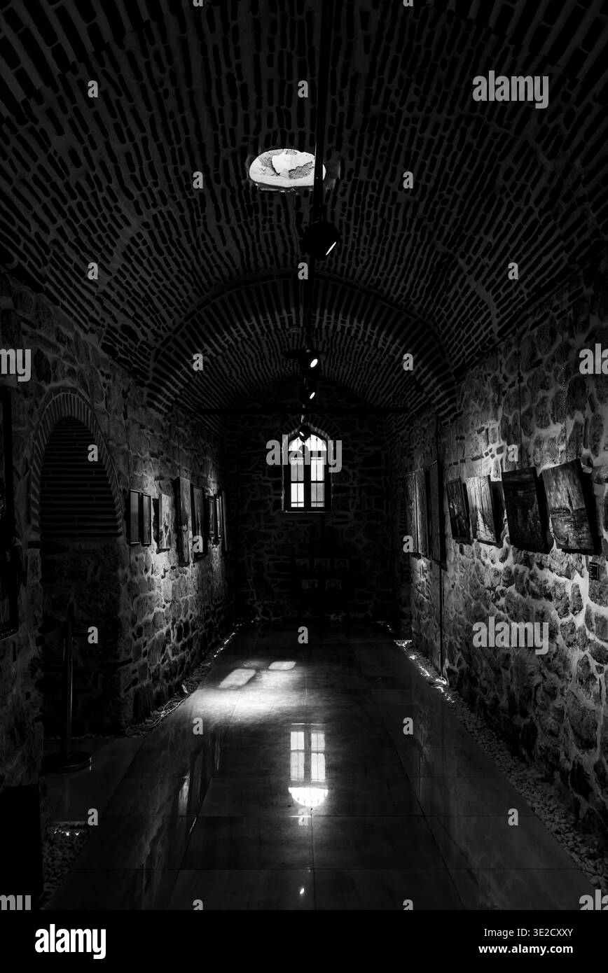Interni in bianco e nero moodici di uno stretto corridoio storico in pietra con soffitto ad arco in mattoni incorniciati d'arte sulle pareti e una luminosa finestra alla fine Foto Stock