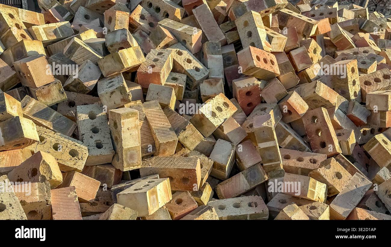 Un enorme e caotico mucchio di mattoni di argilla perforati in tonalità calde di abbronzatura e arancio. I blocchi sparsi proiettano ombre profonde e nitide sotto la luce del sole. Foto Stock