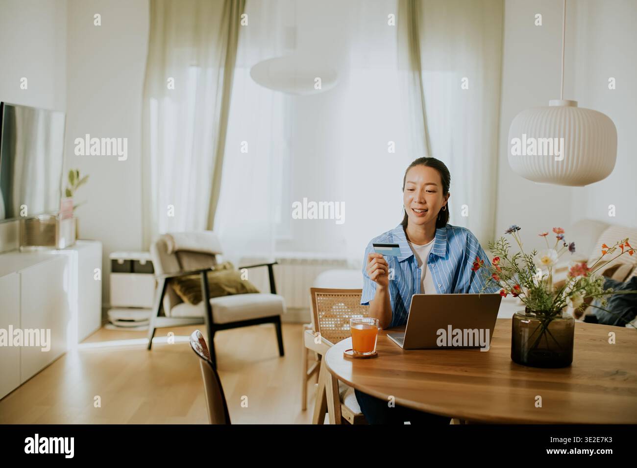 Una donna siede su un tavolo di legno con un portatile aperto davanti a lei. Sta sorridendo mentre ha una carta. La luce del sole riempie la stanza e ci sono piante Foto Stock