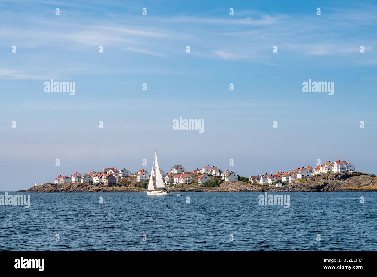 Barca a vela che passa per l'isola di Bohuslän a Åstol, costa occidentale della Svezia, con case bianche sul lungomare sulla costa rocciosa sotto il cielo blu Foto Stock