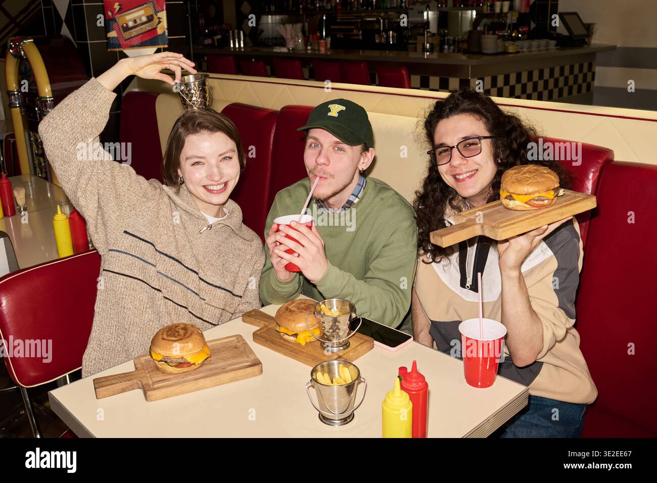 Ritratto di tre allegri studenti della gen Z che sorridono per la macchina fotografica gustando hamburger e bevande al ristorante retrò Foto Stock