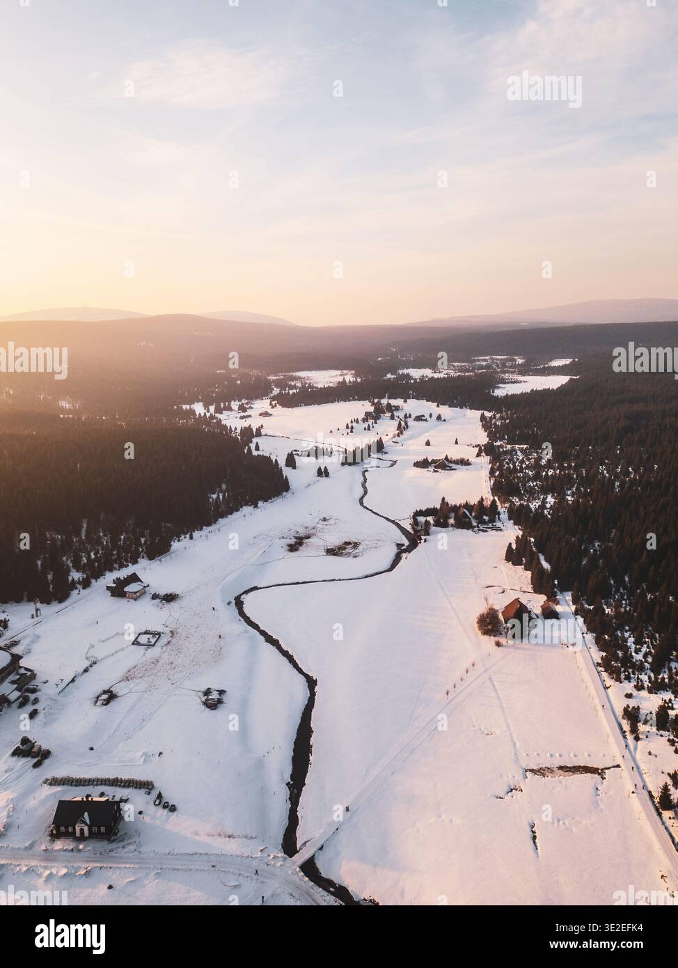 La neve copre il villaggio di Jizerka, circondato da alberi e colline. Il sole tramonta, illuminando il fiume tortuoso e i campi aperti, creando una scena invernale in montagna. Foto Stock