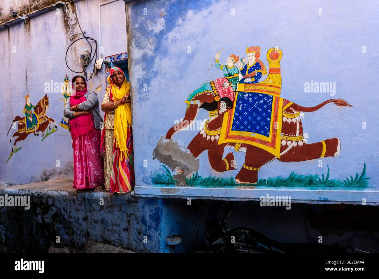 Due donne che indossano il saris si trovano in una porta a Bundi accanto a un grande e colorato murale raffigurante una figura reale che cavalca un elefante decorato. Foto Stock