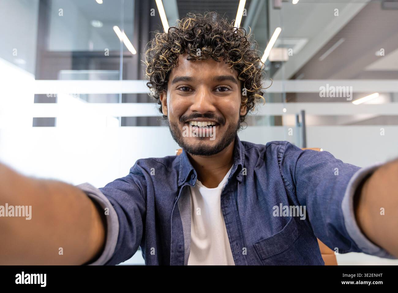 Ritratto ravvicinato di un giovane indiano in un ufficio, sorridente e che parla con la macchina fotografica che ha in mano. Foto Stock