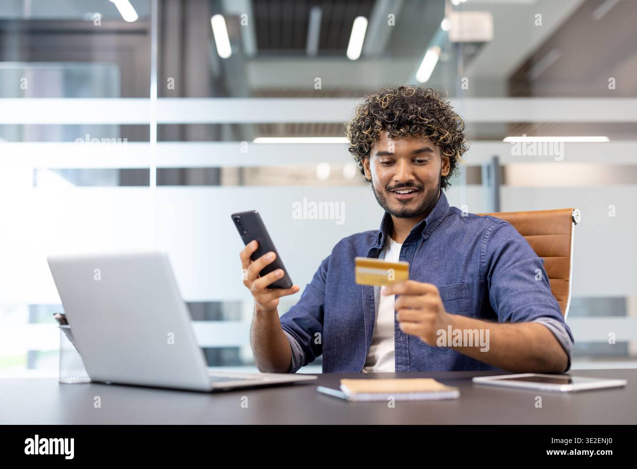 Sorridente giovane indiano seduto alla scrivania dell'ufficio, utilizzando il telefono cellulare e la carta di credito. Foto Stock